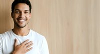 Young man with dark hair and beard, South Asian ethnicity, wearing white casual t-shirt, hand on chest, smiling warmly against wooden background. Portrait for business, healthcare, wellness, gratitude themes.