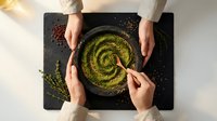 Green smoothie bowl with spirulina powder being eaten by two people sharing healthy breakfast meal from above on dark slate board.