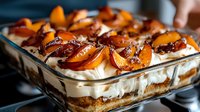 Layered dessert with caramelized peach slices, whipped cream, and crumbled cookies in a glass baking dish, perfect for bakery menus and food photography.