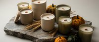 Scented candles in neutral ceramic containers arranged on marble base with dried wheat and decorative gourds for autumn home decor and seasonal styling.