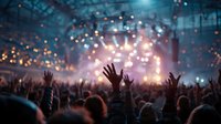 Concert crowd with raised hands enjoying live music performance under bright stage lights and bokeh effects at indoor venue.