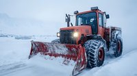 Red snow plow tractor with large blade clearing snowy winter road during heavy snowfall, dramatic mountain landscape in background, cold weather scene.