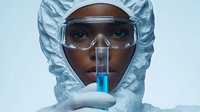 Young African American female scientist in protective suit and goggles holds test tube with blue liquid while conducting laboratory research. Clean medical background.
