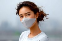 Young Asian woman wearing protective face mask outdoors with wind blowing through hair, urban background with soft bokeh effect, natural lighting.