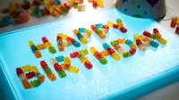 Happy Birthday written with colorful gummy bears on turquoise surface with scattered candies, celebration greeting card design for parties and festive occasions.
