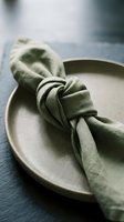 Sage green linen napkin elegantly tied with knot on neutral ceramic plate, styled for fine dining, table setting, hospitality, and event planning concepts.