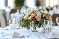 Elegant table setting with peach roses and white floral centerpiece in glass vase, surrounded by wine glasses on crisp white tablecloth for wedding reception.