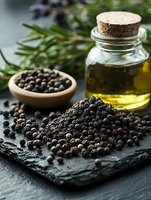 Black peppercorns scattered on dark stone surface with glass bottle of oil, wooden bowl and fresh rosemary sprigs in moody natural lighting.