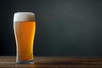 Glass of cold draft beer with foam head and condensation on dark background, wooden table surface with copy space for restaurant menu design.