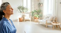 Mature woman with gray hair and medium skin tone wearing blue shirt gazing at bright, minimalist living room with potted plants, wooden furniture and natural light from windows.