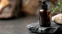 Dark amber glass pump bottle on black stone with water drops, minimal spa composition against blurred natural background for cosmetic product presentation.