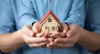 Man in blue shirt holding small wooden house model in cupped hands representing home protection, real estate investment, mortgage insurance concept.