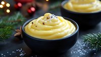 Creamy yellow custard dessert topped with black vanilla seeds in dark ceramic bowl against festive holiday background with pine branches and bokeh lights.