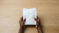 Person with dark skin tone holding folded white t-shirt on wooden surface, displaying garment for retail, fashion merchandising, or clothing business purposes.