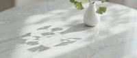 Young woman with fair complexion arranging fresh green parsley in white ceramic vase on marble table with natural sunlight casting leaf shadows for home decor and lifestyle content.