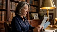 Mature woman with shoulder-length gray hair, wearing dark navy blouse, seated at wooden desk in home library, holding tablet device, surrounded by bookshelves and brass lamp, professional setting.