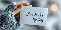 Female hand holding white card with handwritten message "You make my day" against soft bokeh background. Cozy winter sweater creates warm, encouraging mood.