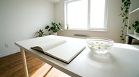 Minimalist home office workspace with open notebook and glass bowl on white desk near window with natural light and green plants.