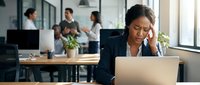 Stressed African American businesswoman experiencing headache while working on laptop in modern office with colleagues discussing in background.