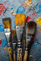 Paint brushes with colorful oil paint strokes on textured gray background, showing artistic tools with vibrant yellow, blue and red paint splatters and smears.