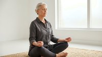 Senior Caucasian woman practicing meditation in lotus pose on yoga mat by window in bright modern room for wellness and mindfulness lifestyle.