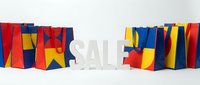 Colorful red, blue, and yellow shopping bags arranged around a white SALE sign on a light background, perfect for retail promotions and seasonal discount campaigns.