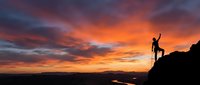 Silhouette of triumphant hiker with raised arms celebrating success on mountain peak against dramatic orange sunset sky. Achievement and adventure concept.