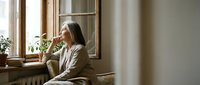 Mature woman with long dark hair and light skin tone, wearing a beige cardigan, sitting by a window in contemplative pose, suitable for wellness, mental health, lifestyle, and editorial content.