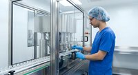 Male pharmaceutical technician in blue scrubs and hairnet operating sterile manufacturing equipment in modern cleanroom facility for drug production.