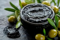 Black natural charcoal face mask in glass jar with fresh green olives and leaves on dark stone background, skincare and spa treatment concept.