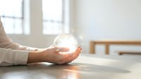 Female hands holding glowing crystal ball on wooden table with bright window light, symbolizing future vision, spirituality, and mystical energy for wellness and divination concepts.
