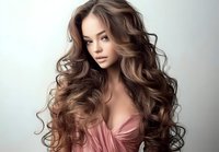 Young woman with long wavy brown hair and pink dress posing against light background for beauty and fashion photography.