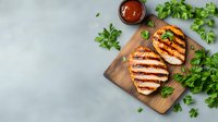 Grilled chicken breast slices on wooden cutting board with fresh parsley and barbecue sauce, top view on light gray background. Food photography.