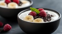 Creamy yogurt bowl topped with fresh raspberries, sliced bananas, dark chocolate chips and mint leaves, served in black ceramic bowl against dark background.