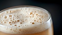 Creamy foam bubbles on top of fresh beer in glass against dark background, macro close-up shot showing texture and carbonation details.
