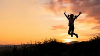 Silhouette of person jumping with arms raised against golden sunset sky over rural landscape, expressing freedom and joy.