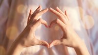 Hands forming a heart shape gesture with warm bokeh light background, symbolizing love, care, and romance for Valentine's Day.