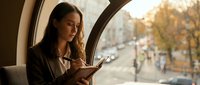 Young professional woman using smartphone while traveling by train, looking out window at autumn city street during golden hour commute.