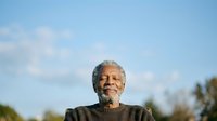 Senior man with gray beard and dark hair, African descent, wearing olive sweater, outdoors against blue sky with clouds and blurred foliage background for portrait and lifestyle use.