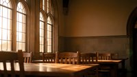 Empty wooden tables and chairs in warm sunlit room with large arched windows creating peaceful study or meeting space for educational institutions.