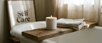 Rustic wooden bathtub tray with a lit candle and linen towel, self care sign in background, cozy spa bathroom atmosphere.