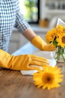 Person wearing yellow rubber gloves wiping wooden table with white cloth next to glass vase of fresh sunflowers. Casual home cleaning scene with natural light and bokeh background.