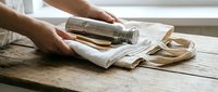 Hands wrapping glass jar with white paper on rustic wooden table for eco-friendly packaging and sustainable gift presentation in natural light setting.