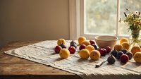 Fresh colorful stone fruits peaches and plums scattered on linen cloth near window with natural sunlight creating warm kitchen atmosphere for cooking.