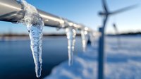 Frozen icicles hanging from metal pipes at wind turbine farm on winter day, blurred windmill silhouette in background against blue sky and snowy landscape.