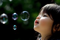 Young Asian child with dark hair looking up in wonder at floating soap bubbles outdoors with natural green bokeh background capturing pure joy and childhood innocence.