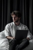 Young Caucasian man with curly hair working on laptop in bed wearing white shirt against dark curtain background for remote work and lifestyle concepts.