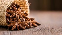 Star anise pods spilling from rustic burlap sack onto wooden surface, aromatic spice composition with warm brown tones and selective focus creating depth.