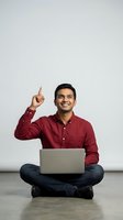 Young South Asian male professional wearing maroon button-up shirt and dark jeans, seated cross-legged with laptop, pointing upward with confident smile against white background for business and technology concepts.