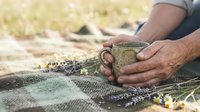 Senior man hands holding rustic ceramic mug outdoors on woven blanket with wildflowers for cozy camping morning coffee ritual and peaceful nature moments.